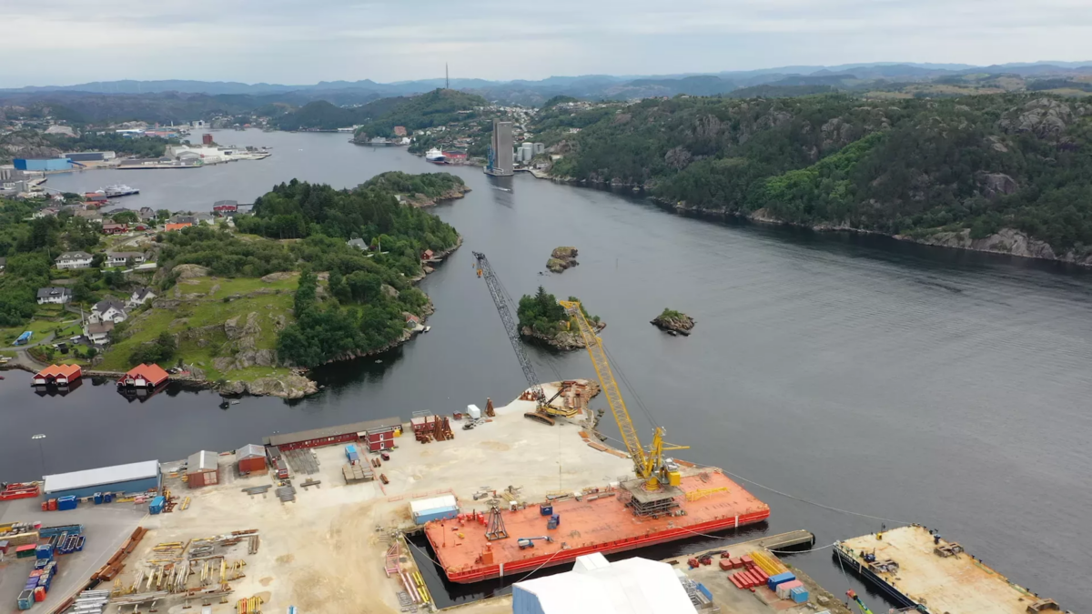 Photograph in eagle perspective showing a outdoor testing area with a large crane in focus. The crane is positioned on a platform floating on water (a river).