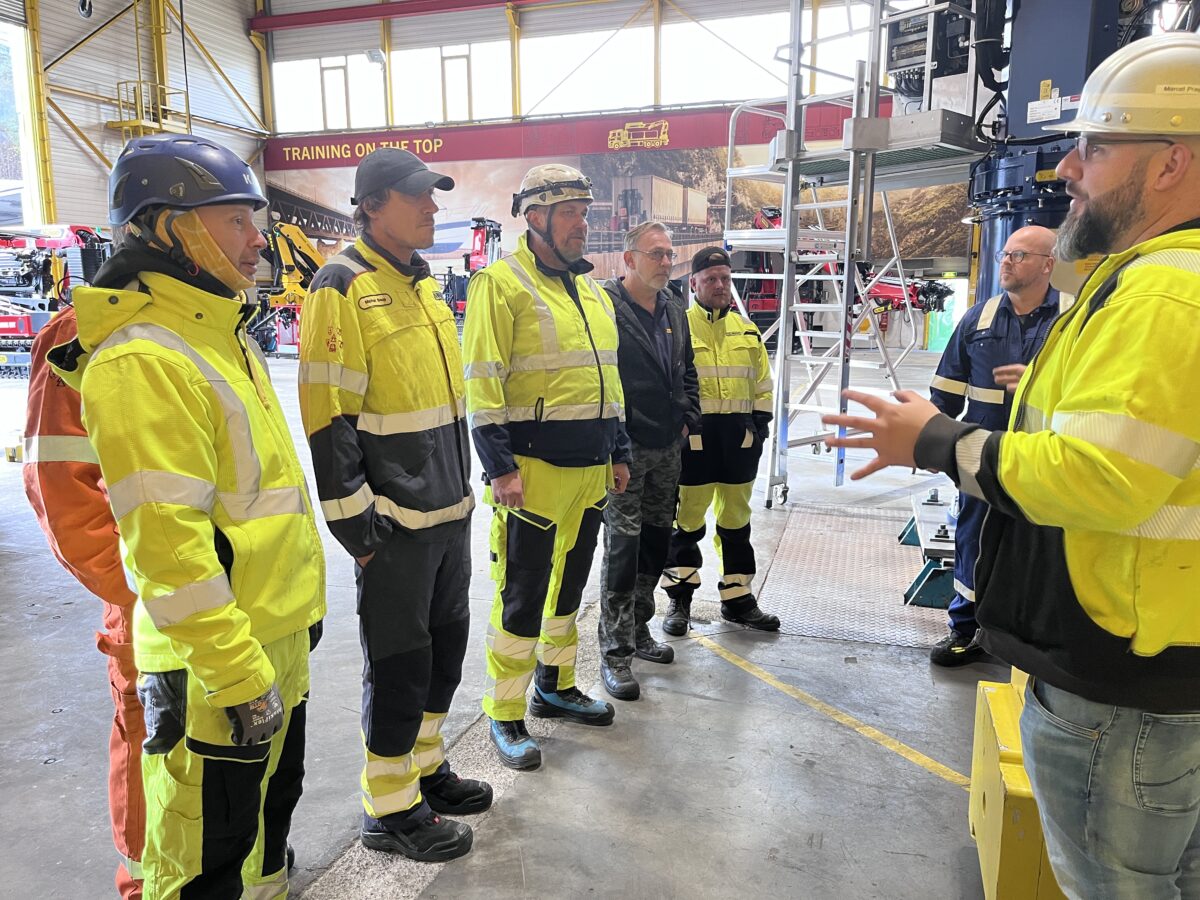 Six training attendees standing in a row in a training center, wearing protective clothing, listening to a training instructor, who also wers protective clothing.