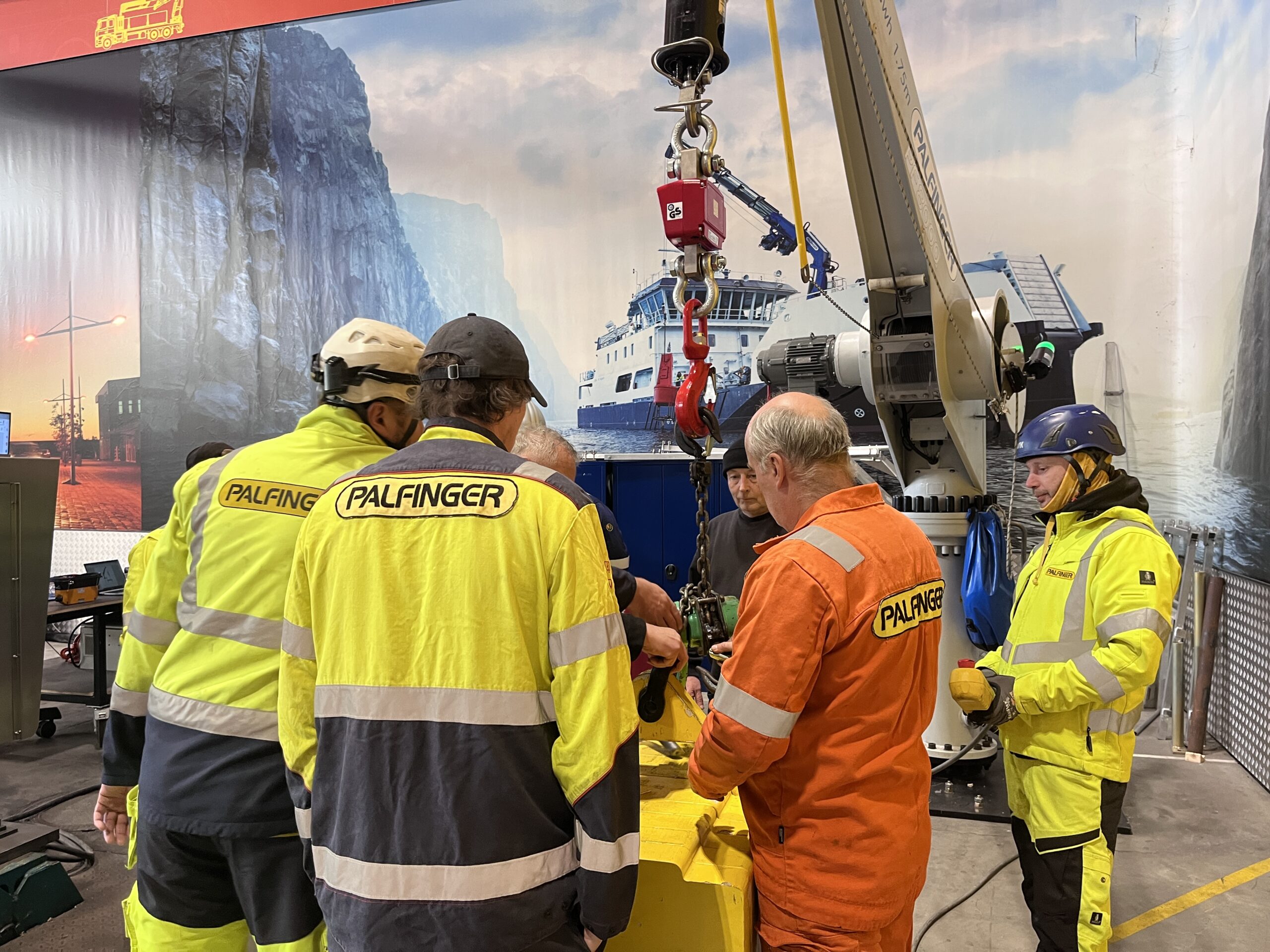 Training attendees standing in a circle around a heavy yellow object, which serves as a weight for testing and training purposes. The weight is attached to the hook of a crane, which is located in the background, behind the group.