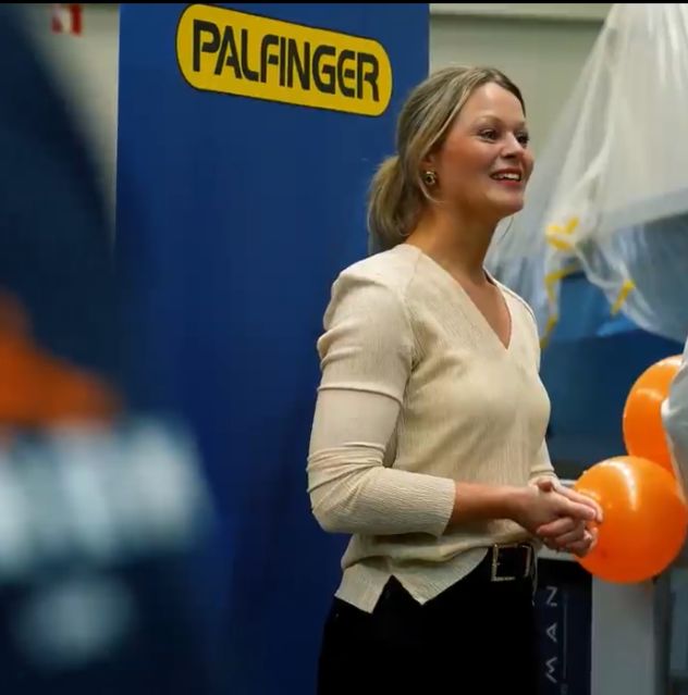 Woman standing in front of a blue roll-up with PALFINGER logo. She is presenting and smiling at the audience, who is not visible in the photograph.