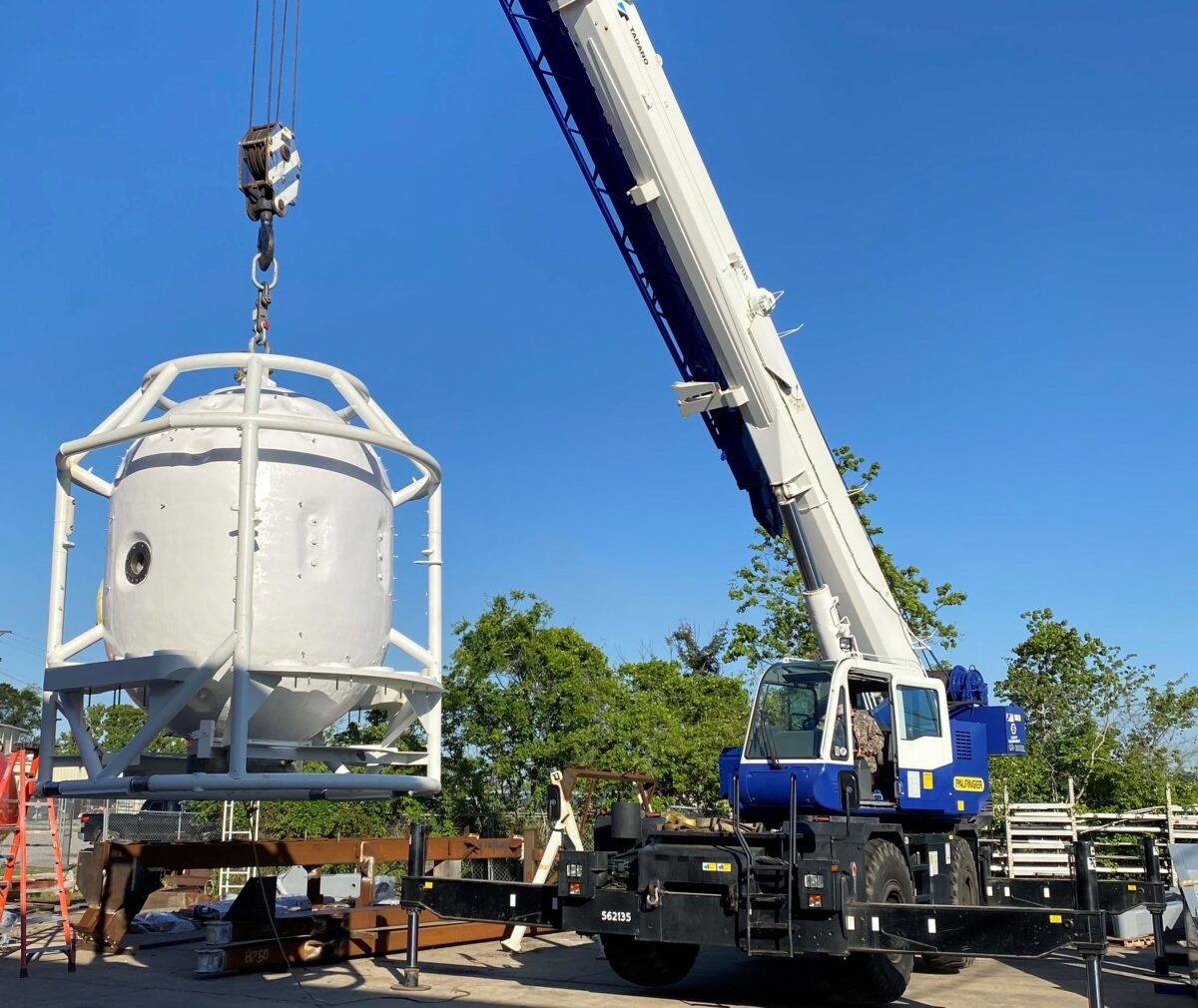 A crane hoisting a diving bell, which has the shape of a capsule and an opening for divers to enter and exit.