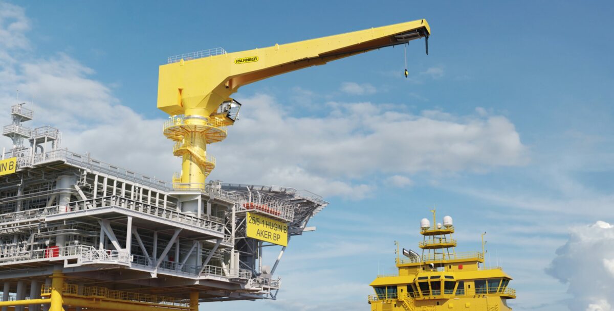 Big yellow crane mounted on an offshore platform, prepared to lift materials from a vessel onto the platform. The vessel is slightly visible in the right bottom corner of the image.