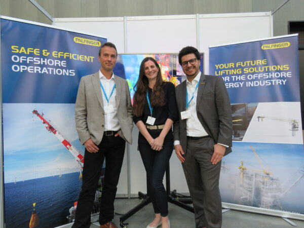 Three people (two men and one woman) standing in front of PALFINGER MARINE branded roll-ups, smiling into the camera. They are attending the Offshore Crane and Lifting Conference in Stavanger, Norway.