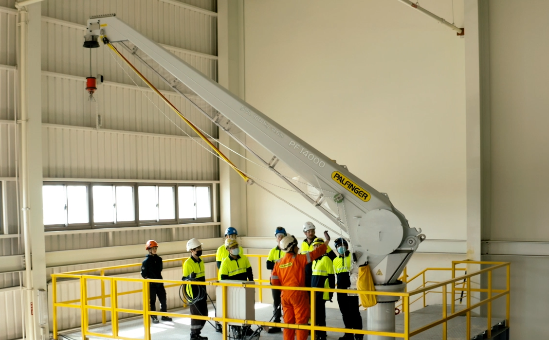 PALFINGER fixed boom crane in a hall, surrounded by a couple of training participants and one trainer clad in an orange, PALFINGER branded, overall. The trainer, looking and gesturing at the crane, is talking to the attendees.