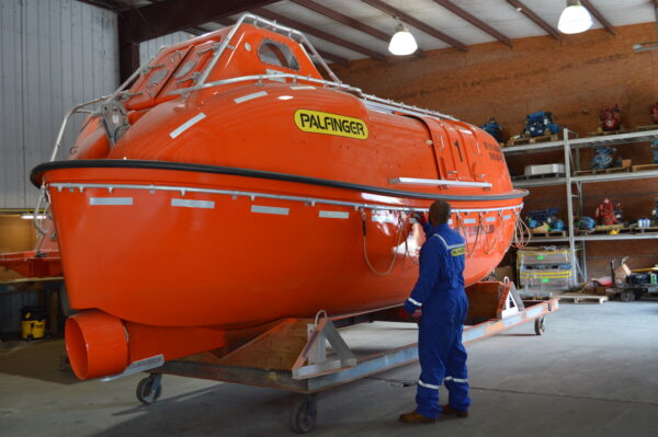 A service engineer in a workshop doing a refurbishment job on an orange, totally enclosed lifeboat.