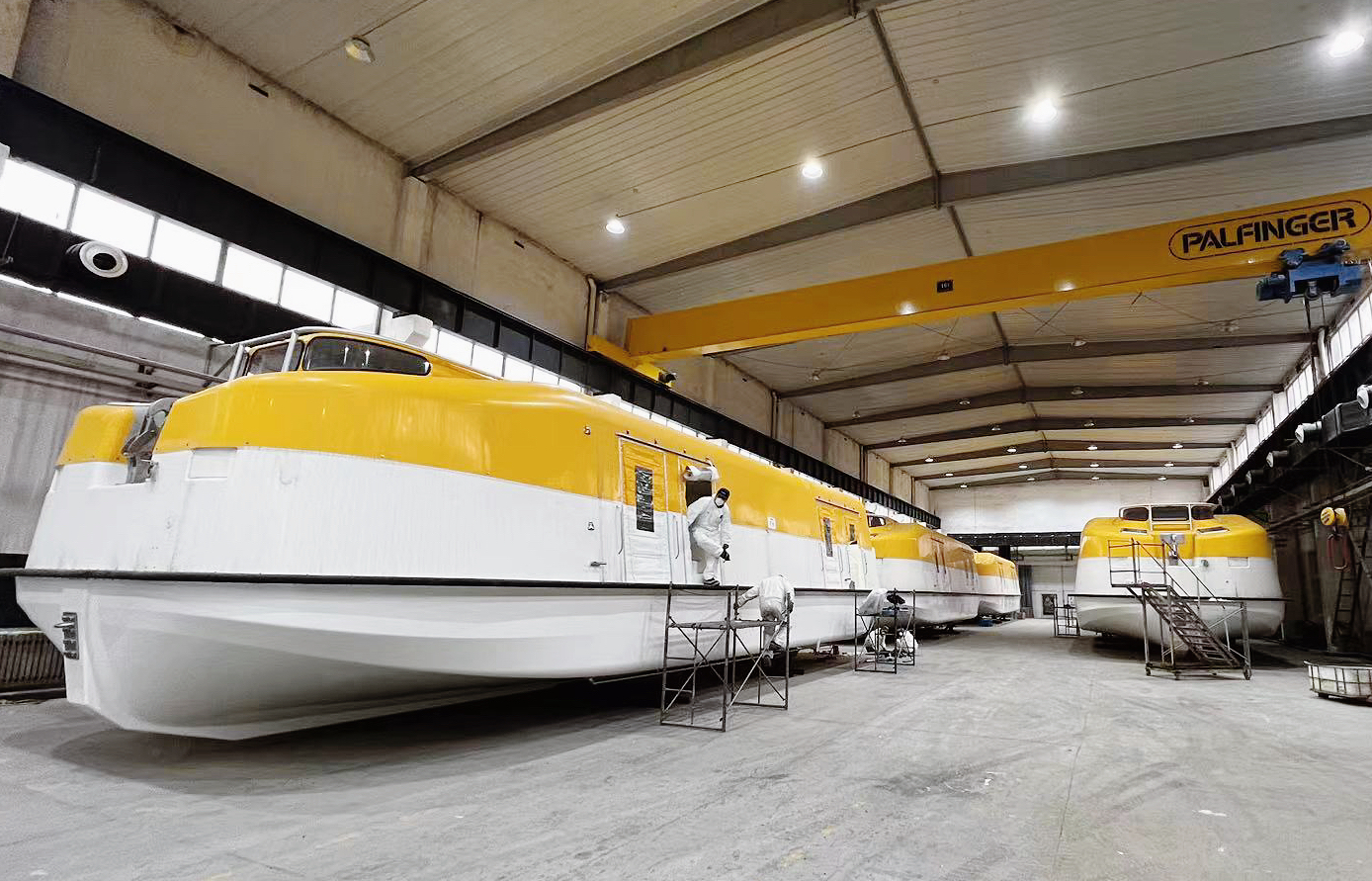 Four totally enclosed lifeboats in a production hall. Four people clad in white work gear are servicing the boats.