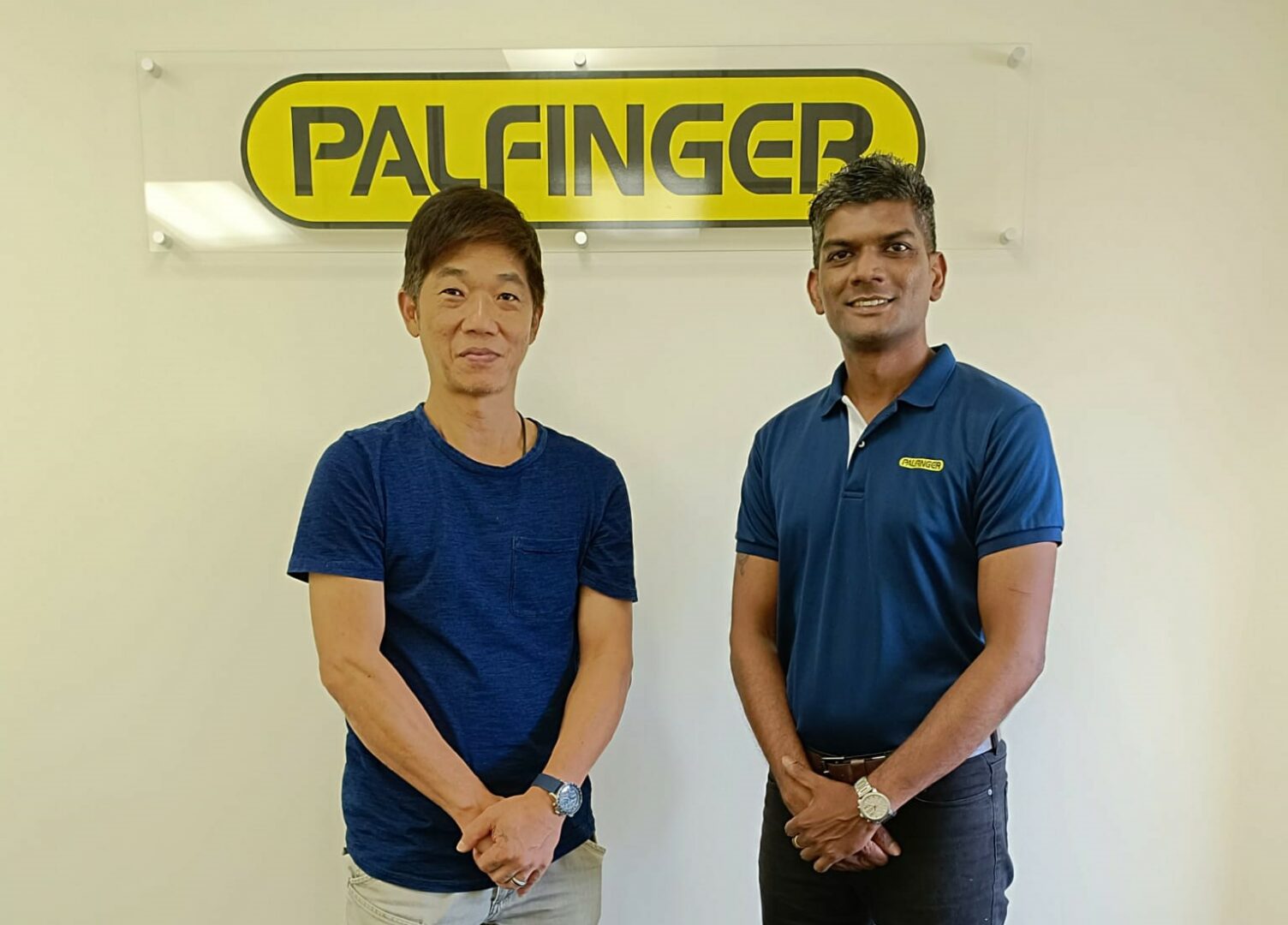 Two men - both wearing blue shirts - standing in front of a wall (with a PALFINGER logo attached to it), smiling into the camera.