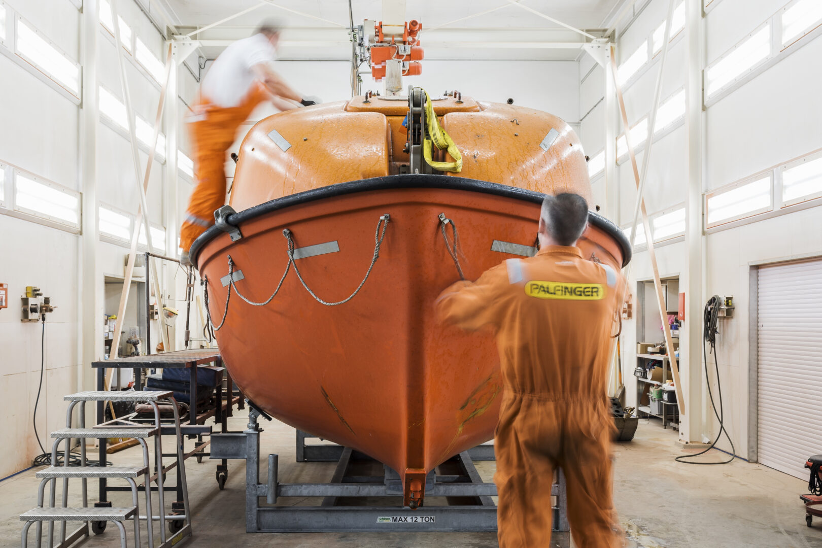 Two man clad in workwear in a workshop doing maintenance/service work on an orange colored, totally enclosed lifeboat.